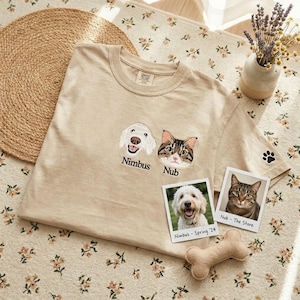 May include: A beige short-sleeve t-shirt featuring embroidered portraits of a white dog and a brown tabby cat with the names Nimbus and Nub stitched below. A small black paw print is embroidered on the sleeve, accompanied by two polaroid-style photos.