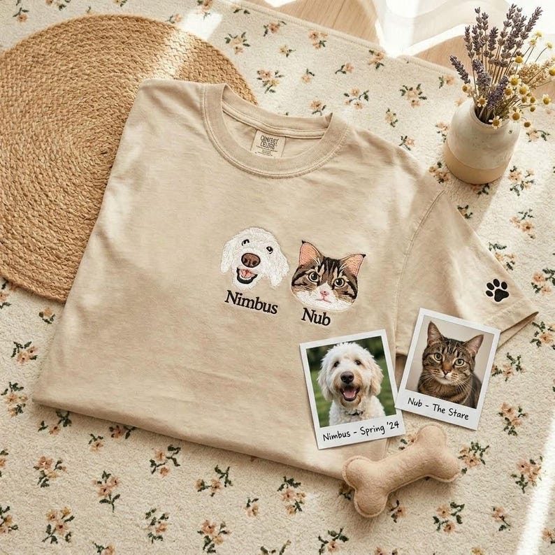 May include: A beige short-sleeve t-shirt featuring embroidered portraits of a white dog and a brown tabby cat with the names Nimbus and Nub stitched below. A small black paw print is embroidered on the sleeve, accompanied by two polaroid-style photos.