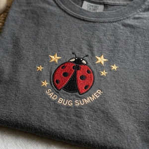May include: A charcoal gray t-shirt featuring an embroidered design of a red and black ladybug surrounded by small yellow stars. Below the ladybug, the text SAD BUG SUMMER is embroidered in yellow thread. The shirt has a Comfort Colors label.
