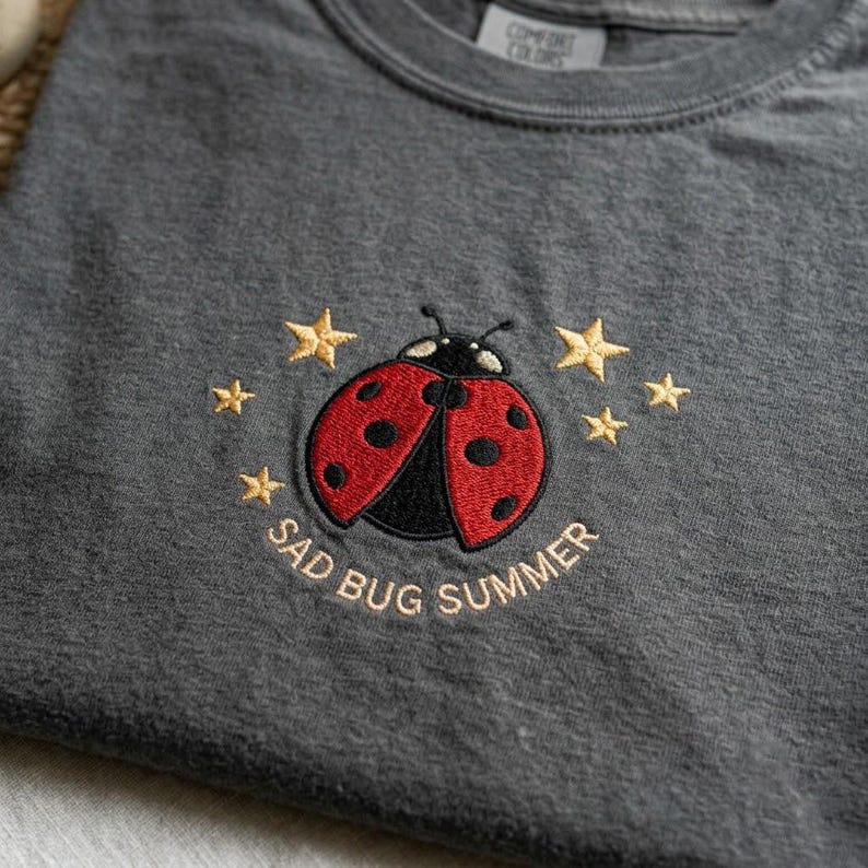 May include: A charcoal gray t-shirt featuring an embroidered design of a red and black ladybug surrounded by small yellow stars. Below the ladybug, the text SAD BUG SUMMER is embroidered in yellow thread. The shirt has a Comfort Colors label.