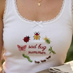 May include: A white ribbed tank top with lace trim along the neckline and a small white bow. The center features embroidered insects including a ladybug, beetle, butterfly, caterpillar, and ants, with the red text "sad bug summer" in the middle.