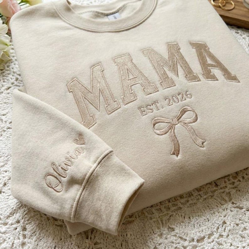 Peut inclure: Un sweat-shirt beige avec le mot "MAMA" brod&eacute; en marron clair, dans une police de style universitaire. En dessous du mot "MAMA" se trouve "EST. 2026" et un n&oelig;ud. Le poignet de la manche gauche porte le nom "Olivia" brod&eacute; avec un c&oelig;ur.