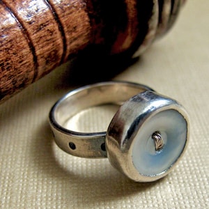 May include: A silver ring with a white button set into the top. The button has a silver thread running through the center.