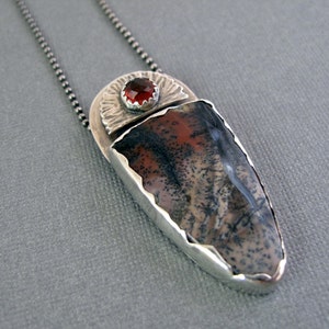 May include: A silver necklace with a pendant featuring a red gemstone surrounded by a silver sunburst design. The pendant is set in a silver frame with a jagged edge and a brown and gray agate stone.