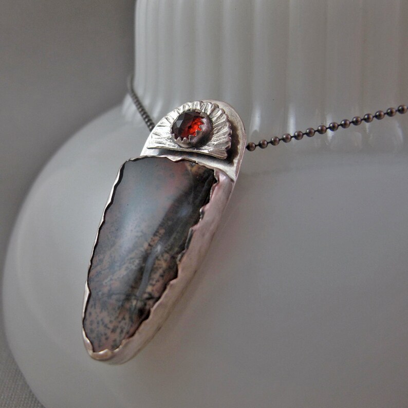 May include: A silver pendant necklace with a dark gray and brown agate stone and a red gemstone set in a silver sunburst design.