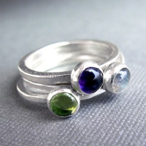 Puede incluir: Tres anillos de plata con piedras preciosas de diferentes colores. Los anillos están apilados uno encima del otro. El anillo superior tiene una amatista púrpura, el anillo del medio tiene un peridoto verde y el anillo inferior tiene una piedra lunar blanca.
