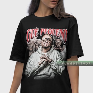 Op de afbeelding: Zwart T-shirt met een grafische print van een man met een baard die een witte hoodie draagt. De tekst "QUE PEQUENO" is in rood op het shirt gedrukt.