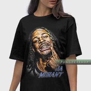 May include: Black t-shirt featuring a graphic of a smiling person with gold teeth, pointing at their mouth. The text "JA MORANT" is displayed in blue and white. The shirt has short sleeves.
