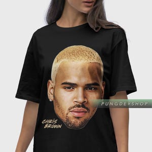 Puede incluir: Camiseta negra con un gran gr&aacute;fico del rostro de Chris Brown. La imagen muestra un primer plano de su rostro con cabello rubio y barba. Las palabras "CHRIS BROWN" est&aacute;n impresas en oro debajo.