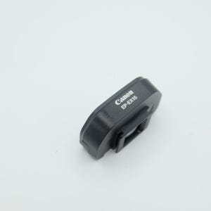 May include: A black Canon EP-EX15 eyepiece extender accessory for a camera. The device features a rectangular mounting frame with a central opening and the brand name Canon and model number EP-EX15 printed in white text on the top surface.