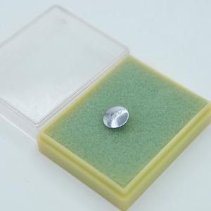 May include: A small, silver-colored, circular object with the word "Topcon" printed on it, resting on a green foam insert inside a clear plastic container with a yellow border. The object has a ridged edge.