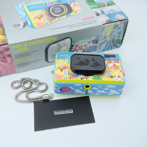 Puede incluir: Cámara Lomography con objetivo gran angular de 35 mm. La cámara tiene un cuerpo azul y amarillo con un diseño lúdico, una parte superior negra y una cadena plateada. También se incluye una tarjeta negra con la palabra "lomography".