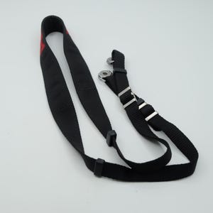 May include: A black nylon camera strap featuring a woven texture, adjustable silver-toned metal buckles, and two circular metal attachment points. A portion of red text is visible on the upper section of the strap, which is looped.