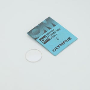 May include: A clear, circular lens next to a blue and white Olympus package. The package reads "OM SYSTEM DIOPTRIC CORRECTION LENS 1 0". The lens is a small, transparent disc.
