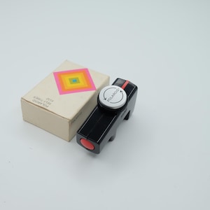 May include: A black Polaroid self-timer #132 device with a red button and a silver dial featuring circular arrows and the text POLAROID. Beside it sits a white cardboard box with a colorful square graphic and the text Polaroid SELF-TIMER #132.