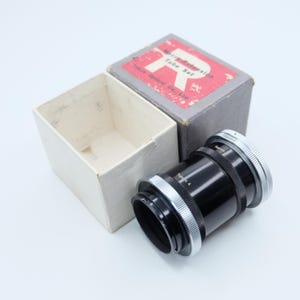 Può includere: Un obiettivo per fotocamera nero e argento con il testo "Topcon 4" e "Teeon Z". L'obiettivo è accanto a una scatola bianca sporca aperta e a una scatola grigia con testo rosso "Macro Extension Tube Set".