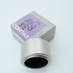 May include: A silver-colored metal lens hood with a black interior, next to its original box. The box is light gray with the text "METAL HOOD GG-3" and the brand name "CONTAX" in white and black.