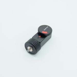 Puede incluir: Accesorio de cámara Polaroid negro con una flecha roja y el nombre de la marca "POLAROID" impreso en la parte superior. El accesorio tiene un dial estriado y un tornillo plateado. El artículo está sobre un fondo blanco.