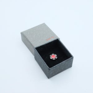 May include: A small, silver-coloured pin with a red centre, displayed in an open grey box with a black interior. The pin has a geometric design, and the box is rectangular.