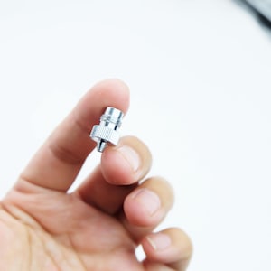 May include: A small, shiny, silver-colored metal screw with a threaded end and a knurled head. The screw is held in a person's hand against a white background. The screw appears to be new and unused.