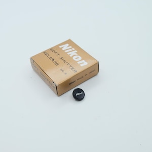 Puede incluir: Un pequeño botón disparador suave negro de Nikon y su caja de cartón color canela. La caja muestra el texto Nikon SOFT SHUTTER RELEASE AR-9 y Nikon MADE IN JAPAN. El botón circular tiene un borde texturizado y el logotipo de Nikon en blanco.