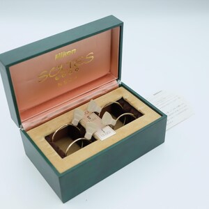 May include: A green box with the brand name "Nikon" and the word "SOLTES" in gold lettering. Inside, there are several gold-colored rings, possibly for a camera lens, and a small tag. The box has a peach-colored interior.