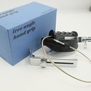 May include: A free angle hand grip with a black leather strap and a silver metal frame. The grip is next to a light blue box with the text "free angle hand grip". The product is designed for photography.
