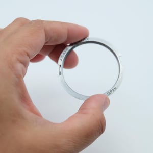 Puede incluir: Un anillo metálico circular de tono plateado con un acabado liso y bordes con muescas. La superficie está grabada con el texto ASAHI y JAPAN en una fuente sans-serif clara. Esta pieza metálica tiene un centro hueco y un perfil plano.
