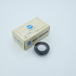 Peut inclure: Un diffuseur plat Minolta et sa boîte d'origine. La boîte est beige avec le logo Minolta et les mots "Flat Diffuser" imprimés dessus. Le diffuseur est noir avec un centre circulaire blanc.