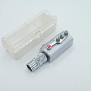 Op de afbeelding: Een zilverkleurig mechanisch apparaat met een rode knop en een doorzichtige plastic behuizing. Het apparaat heeft een cilindrisch lichaam met een schroefdraadeinde en een hendelachtig onderdeel met twee kleine knoppen.