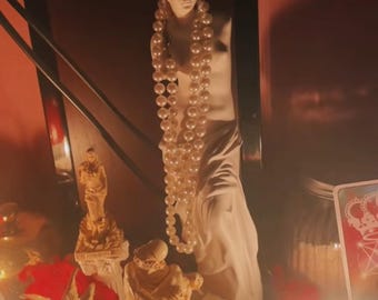 May include: A white statue of a woman draped in fabric, adorned with a long pearl necklace. The scene includes other small statues, red flowers, a lit candle, and a tarot card, creating a mystical ambiance.