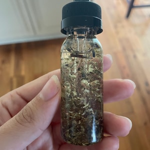 May include: A clear glass bottle with a black dropper top, filled with a liquid and various dried herbs. The bottle is held in a hand, showcasing the contents. The herbs appear to be a mix of green and brown.
