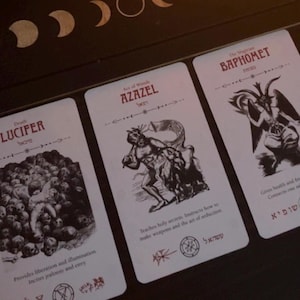 Può includere: Tre carte dei tarocchi con illustrazioni e testo. Le carte presentano le parole "LUCIFER", "AZAZEL" e "BAPHOMET". Le carte hanno illustrazioni di figure e simboli. Lo sfondo è una superficie scura con un disegno a mezzaluna.