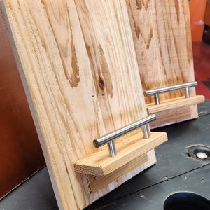 May include: Two wooden phone stands with metal handles. The stands are made of natural wood with a light brown color. The handles are silver and have a simple design.