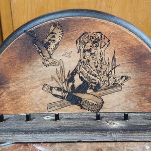 May include: A wooden plaque with a black border in the shape of a half-circle. The plaque features a wood-burned image of a dog with a duck in its mouth, surrounded by other ducks and a hunting scene.
