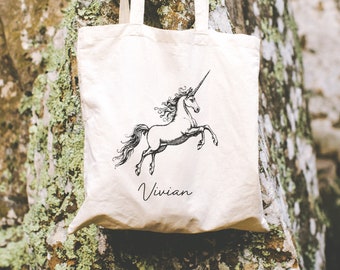 Puede incluir: Una bolsa de lona blanca con una ilustración en blanco y negro de un unicornio y el nombre "Vivian" impreso en la parte delantera.