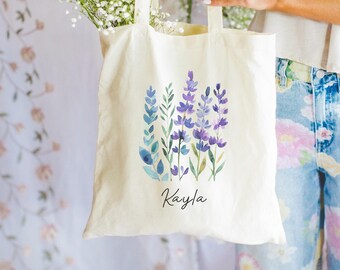 Könnte beinhalten: Eine weiße Canvas-Tragetasche mit einem Aquarell-Design aus lila und blauen Blumen. Die Tasche trägt den Namen "Kayla" auf der Vorderseite.