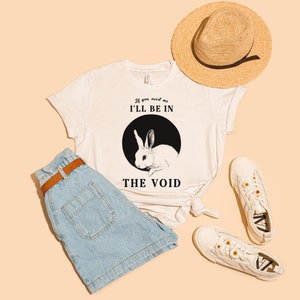 May include: White t-shirt featuring a black and white illustration of a rabbit and the text "If you need me I'll be in the void". The shirt is styled with blue denim shorts, white trainers, and a straw hat.