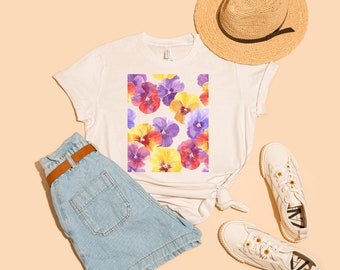 May include: A white t-shirt with a floral print of purple, yellow, and red pansies. The shirt is paired with blue denim shorts, a straw hat, and white sneakers.