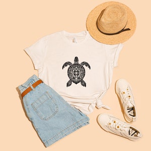 May include: A white t-shirt with a black patterned turtle design. The shirt is paired with blue denim shorts, a straw hat, and white sneakers.