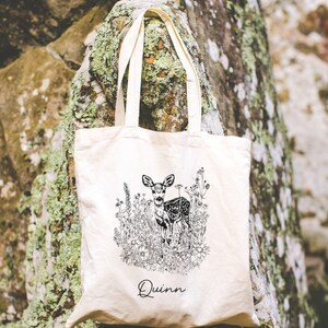 May include: A white canvas tote bag with black and white line art of a deer in a field of flowers. The bag has the name "Quinn" printed on the bottom.