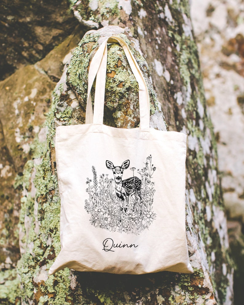 May include: A white canvas tote bag with black and white line art of a deer in a field of flowers. The bag has the name "Quinn" printed on the bottom.