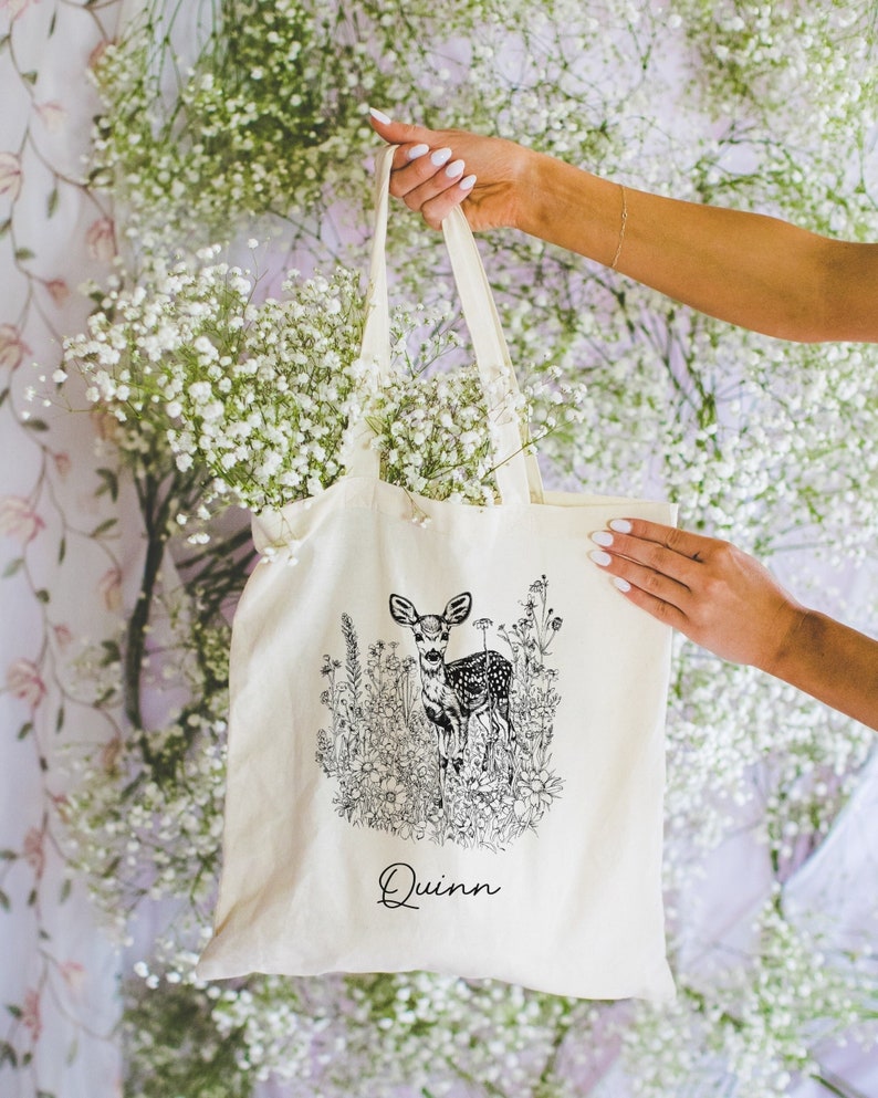 May include: A white canvas tote bag with a black and white illustration of a deer in a field of flowers. The bag has the name "Quinn" printed on the bottom.