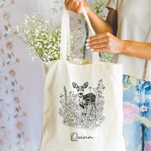 May include: A white canvas tote bag with black and white line art of a deer in a field of flowers. The bag has the name "Quinn" printed on the front.