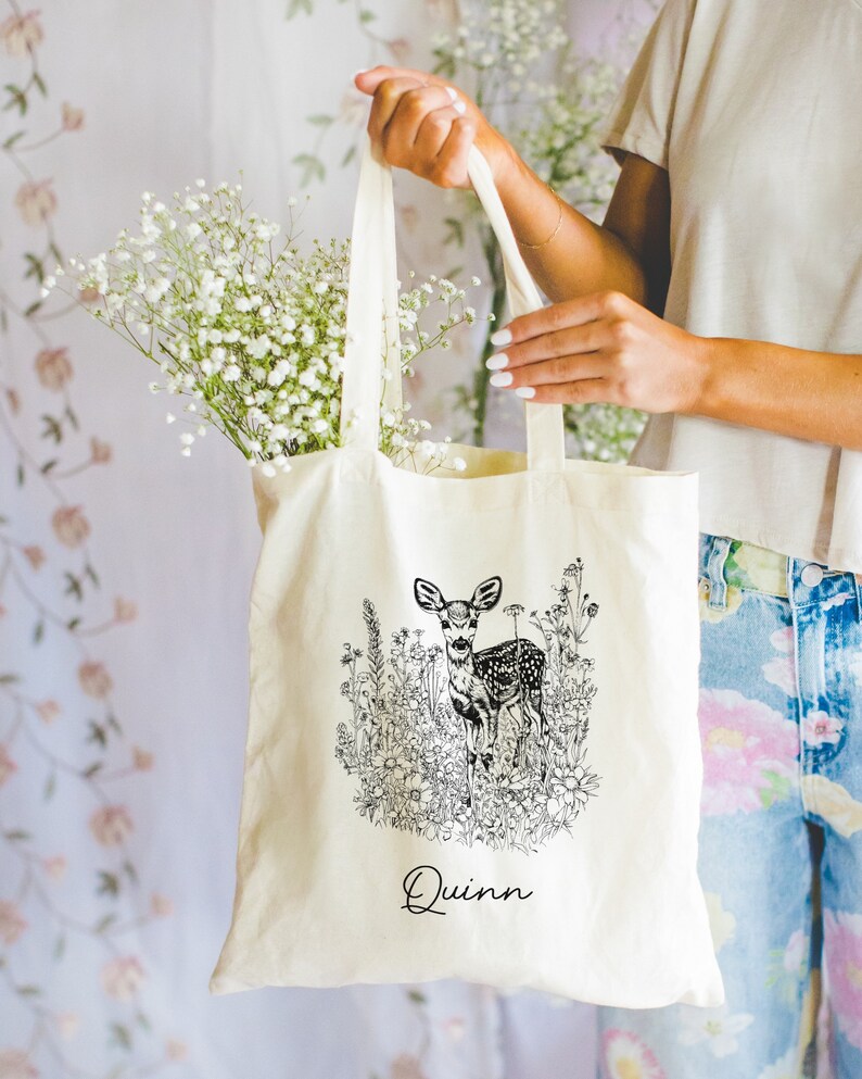 May include: A white canvas tote bag with black and white line art of a deer in a field of flowers. The bag has the name "Quinn" printed on the front.