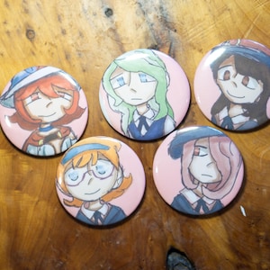 Puede incluir: Cinco botones redondos rosas con ilustraciones de personajes de estilo anime. Cada botón presenta un personaje diferente con diferentes colores de cabello, atuendos y expresiones. Los botones están dispuestos sobre una superficie de madera.