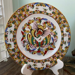 May include: A white porcelain plate with a gold rim and a colourful butterfly design. The butterflies are painted in shades of red, yellow, blue, green, and pink. The plate is on a white stand.