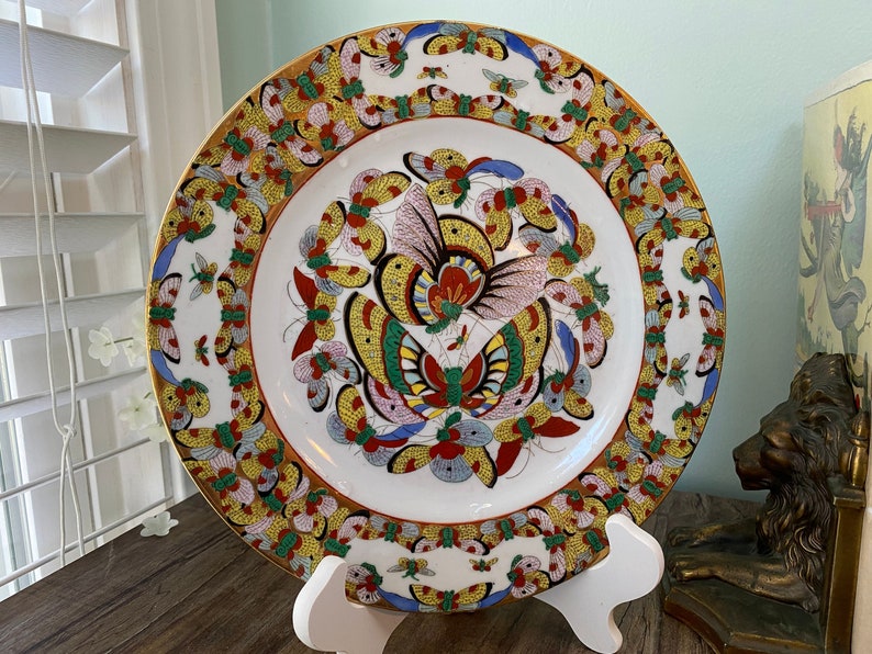 May include: A white porcelain plate with a gold rim and a colourful butterfly design. The butterflies are painted in shades of red, yellow, blue, green, and pink. The plate is on a white stand.