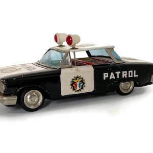 May include: Vintage toy police car in black and white with the word "PATROL" on the door. The car has red lights on top and a colorful emblem on the door. A classic collectible toy.