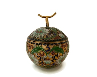 May include: A round, decorative cloisonné box with a lid and a gold-colored handle. The box features intricate floral and foliate designs in green, white, red, and blue against a black and gold background. The overall design is symmetrical and ornate.